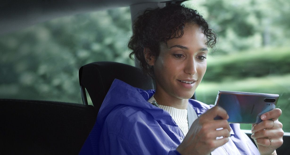 Woman sitting in a car holding Galaxy Note10 plus in landscape mode. Download manager notifications appear showing three large files being downloaded at the same time thanks to the fast data capabilities