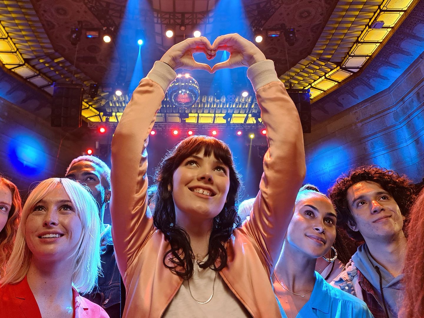 Photo taken with Night mode on Galaxy S20 Ultra of people in a dark venue at a concert, and one women in the middle is making a heart with her hands. The photo is well lit and clear despite the low light environment