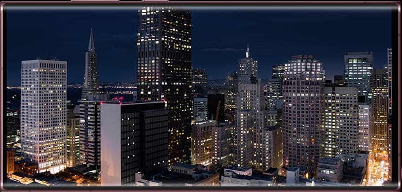 La vue d’un paysage urbain passe de la nuit à la journée. Alors que le ciel s’assombrit pour montrer un ciel nocturne, un zoom arrière s’opère pour afficher une image sur l’écran du Galaxy S22 Ultra. L’accéléré montre toute la durée de la batterie.
