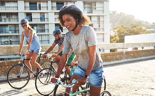 Two women and one man are riding bicycles together on a sunny day. They are similing and enjoying the ride while wearing Galaxy Watch4 devices.