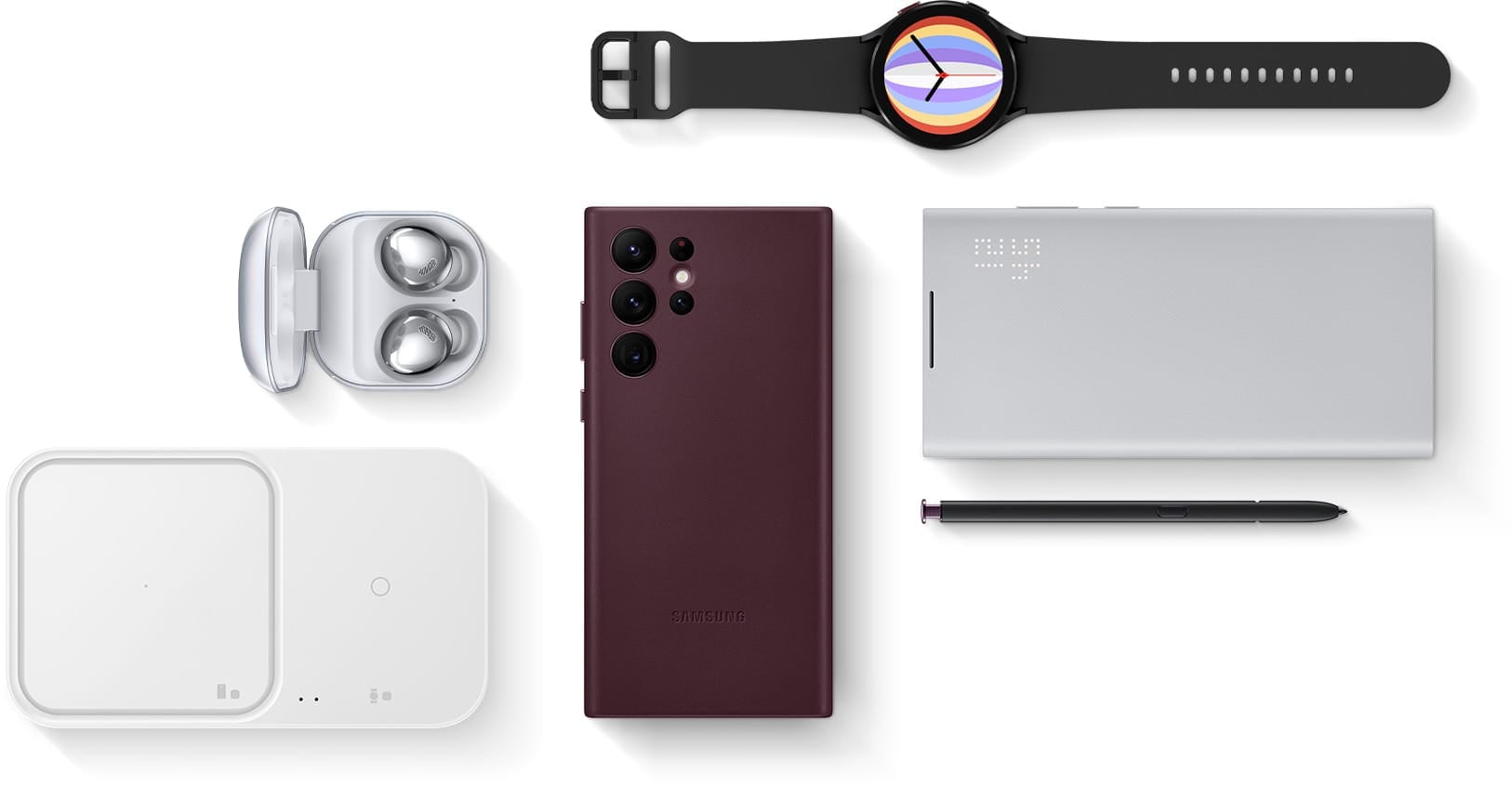 A flat lay of Galaxy Buds Pro with the cradle open and the earbuds in place, Galaxy S22 Ultra seen facedown with the Leather Cover installed, Galaxy Watch4 with an analog graphic on the display, the Super Fast Wireless Charger Duo and Galaxy S22 Ultra seen faceup with the Smart LED View Cover installed with S Pen next to it.