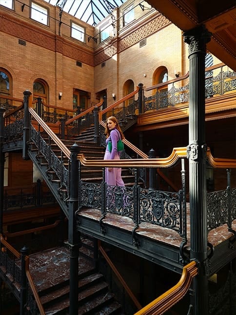 A high resolution photo of a woman standing in a brick atrium with multiple floors of stairs with intricately designed ironwork. Next, the same stairs from another angle and at a distance showing the architecture of the atrium. The 50 megapixel camera is able to capture the scene's rich detail and color. Both photos were taken with Galaxy S23 plus using wide mode with an aperture of F1.8.