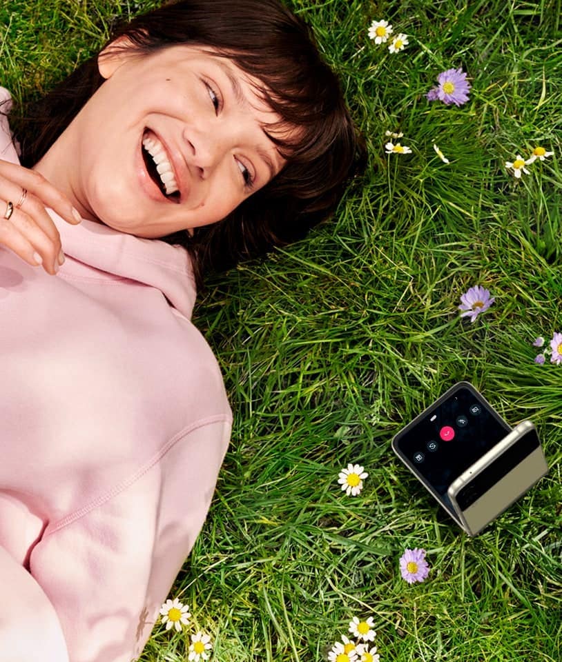 A woman laying in a field, looking into Galaxy Z Flip3 5G in Flex mode.