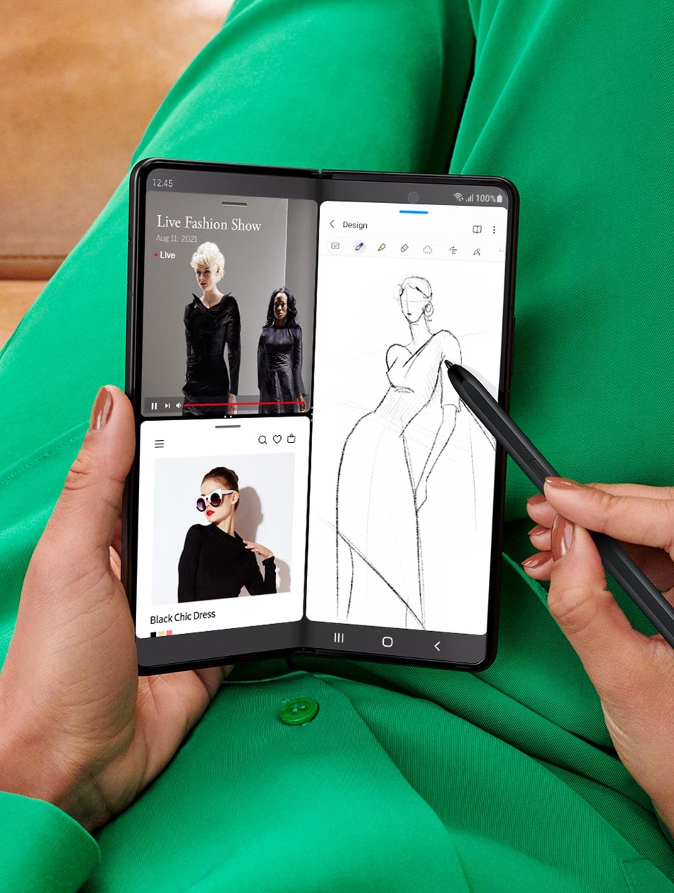 Hands holding unfolded Galaxy Z Fold3 5G and sketching with the S Pen Fold Edition. The Main Screen is in Multi Active Window mode. One half of the screen shows two instances of the Internet app open, one with a live stream of a fashion show and the other with a website showing images of the garments from the show. The other half of the screen is a sketch of a dress being done in Samsung Notes.