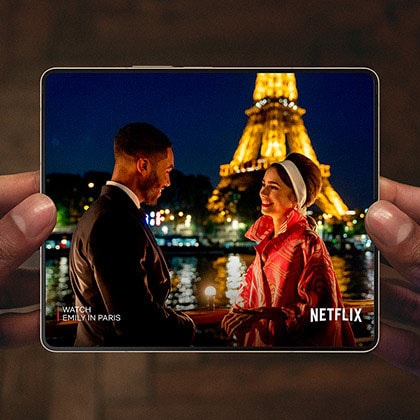 Hands holding an unfolded Galaxy Z Fold4. The Main Screen displays a scene from a Netflix video of a man and woman talking in front of the Eiffel Tower.
