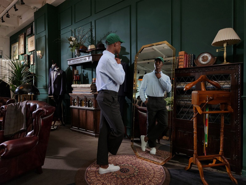 A man stands in a tailor shop and looks at his reflection in a mirror.