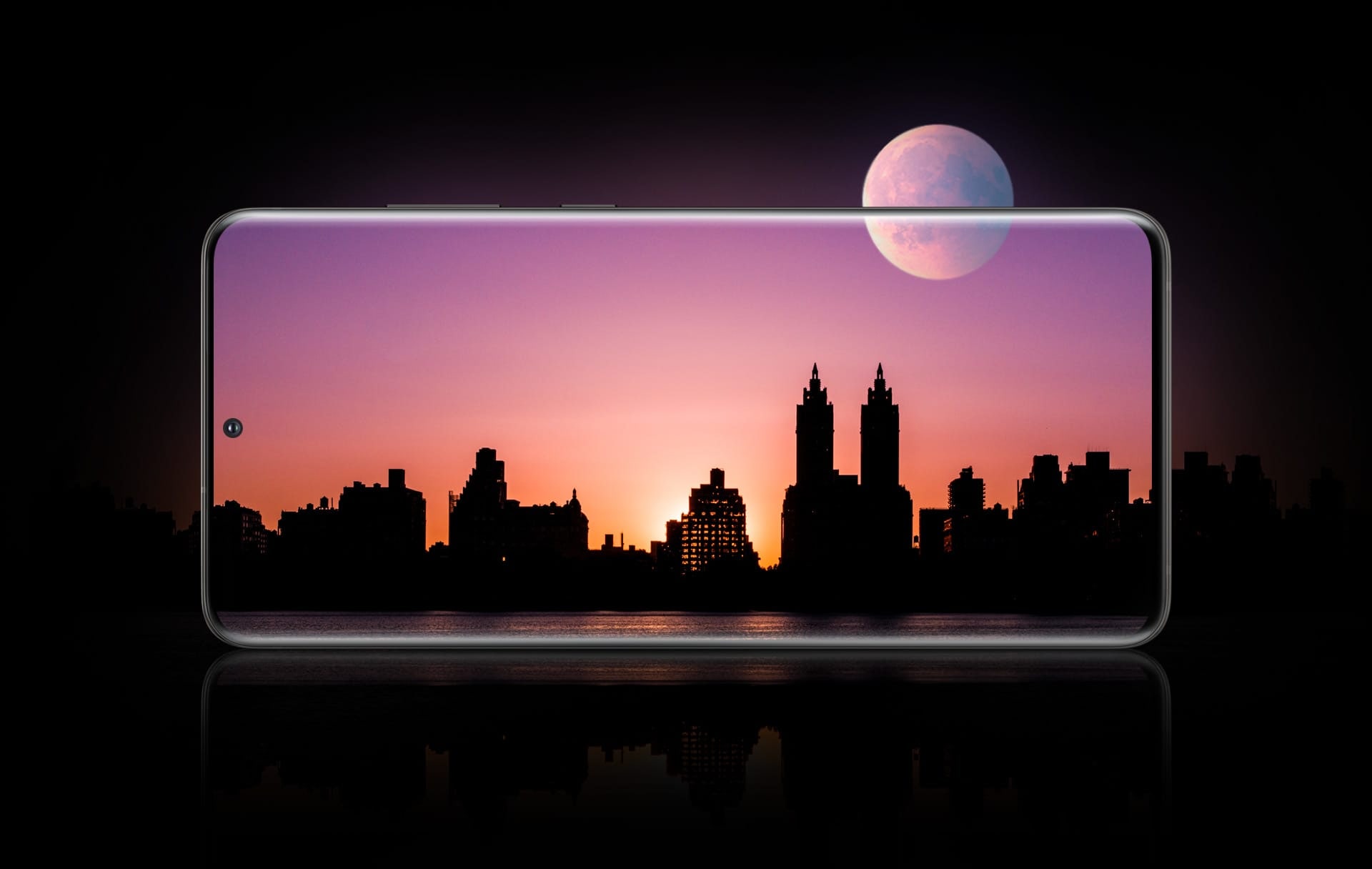 Smartfon Samsung Galaxy S20 Ultra w orientacji poziomej widziany od przodu z nocną panoramą miasta na ekranie. Księżyc na niebie w połowie znajduje się na wyświetlaczu, a w połowie poza nim, obrazuje wytrzymałość baterii dłużej niż jeden cały dzień.