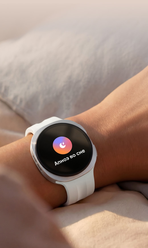 Человек спит в постели с Galaxy Watch9 на руке. На экране отображается функция Отслеживания сна. На лице человека — легкая улыбка, свидетельствующая, что программа Отслеживания сна помогла улучшить его привычки и режим, обеспечив более качественный и восстанавливающий сон.