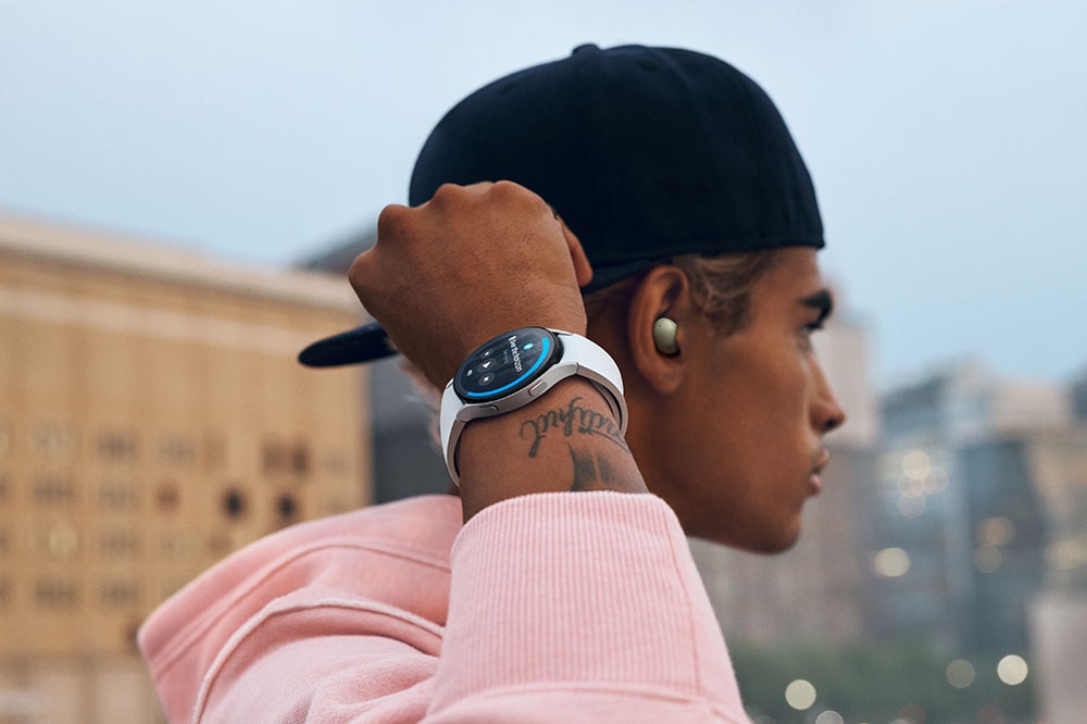 A man with a snapback on backwards is wearing Galaxy Buds2 and a Galaxy Watch4 Classic device. He is holding his right arm up, where he is wearing the watch, and is looking far away in a cityscape.