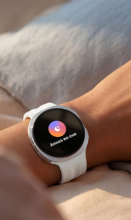 Человек спит в постели с Galaxy Watch9 на руке. На экране отображается функция Отслеживания сна. На лице человека — легкая улыбка, свидетельствующая, что программа Отслеживания сна помога улучшить его привычки и режим, обеспечив более качественный и восстанавливающий сон.