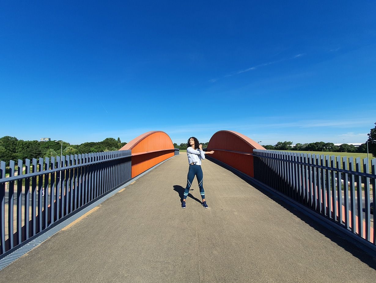 Photo captured by the Ultra Wide Camera of a woman stretching on a bridge with grey gating and orange accents against a bright blue sky
