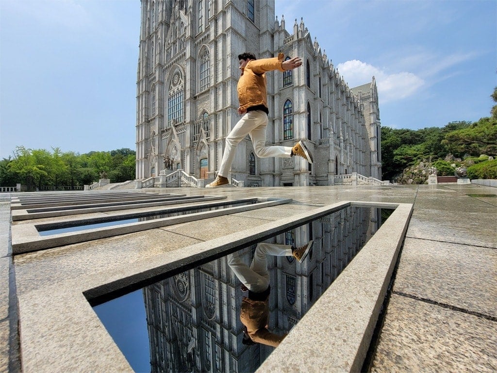 A person jumping across a pool of water in front of a cathedral, taken with the Ultra Wide Camera on Galaxy Note20 Ultra.