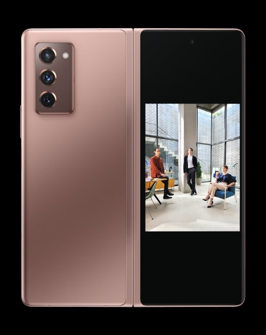 Galaxy Z Fold2 в бронзовом цвете Mystic Bronze в раскрытом виде с интерфейсом приложения камеры и портретом двух мужчин и женщины на основном экране. Телефон разворачивается, показывая основную камеру и внешний экран, на котором виден тот же самый портрет. Таким образом, функция Dual Preview позволяет фотографируемому увидеть кадр еще до того, как будет сделано фото.