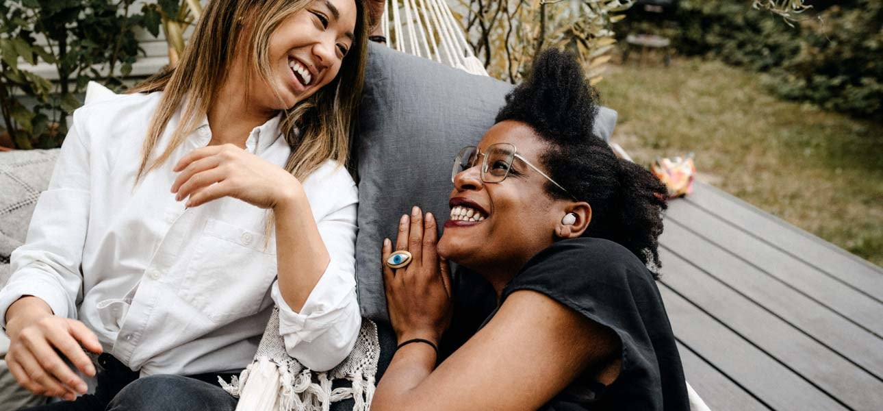 Two women facing each other laughing on a hammock while one of them has white Samsung Galaxy Buds2 Pro in her left ear