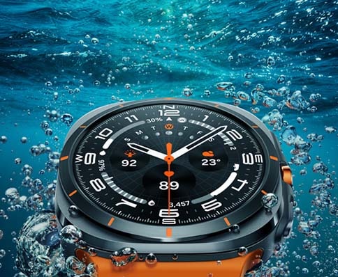 A close-up view of a Samsung smartwatch submerged in clear blue water, surrounded by bubbles. The watch features a dark face with white and orange accents, displaying time, date, and temperature, showcasing its sporty design while promoting.