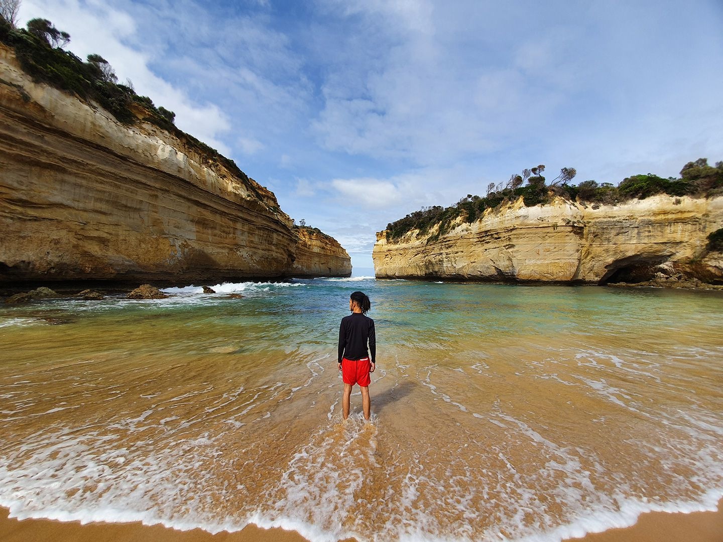 Fotografia zachytená na ultraÅ¡irokouhlý fotoaparát telefónu Galaxy S10 + zobrazujúca muža stojaceho vo vode pred prÃmorskými útesmi.