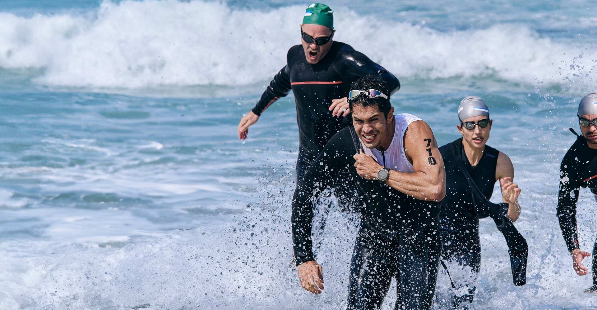 Triatlon atletleri, yüzme sonrası denizden koşarak çıkıyor. Galaxy Watch Ultra takmış bir adam, bir sonraki spora geçiş yapmak için mayosunu çıkarıyor.