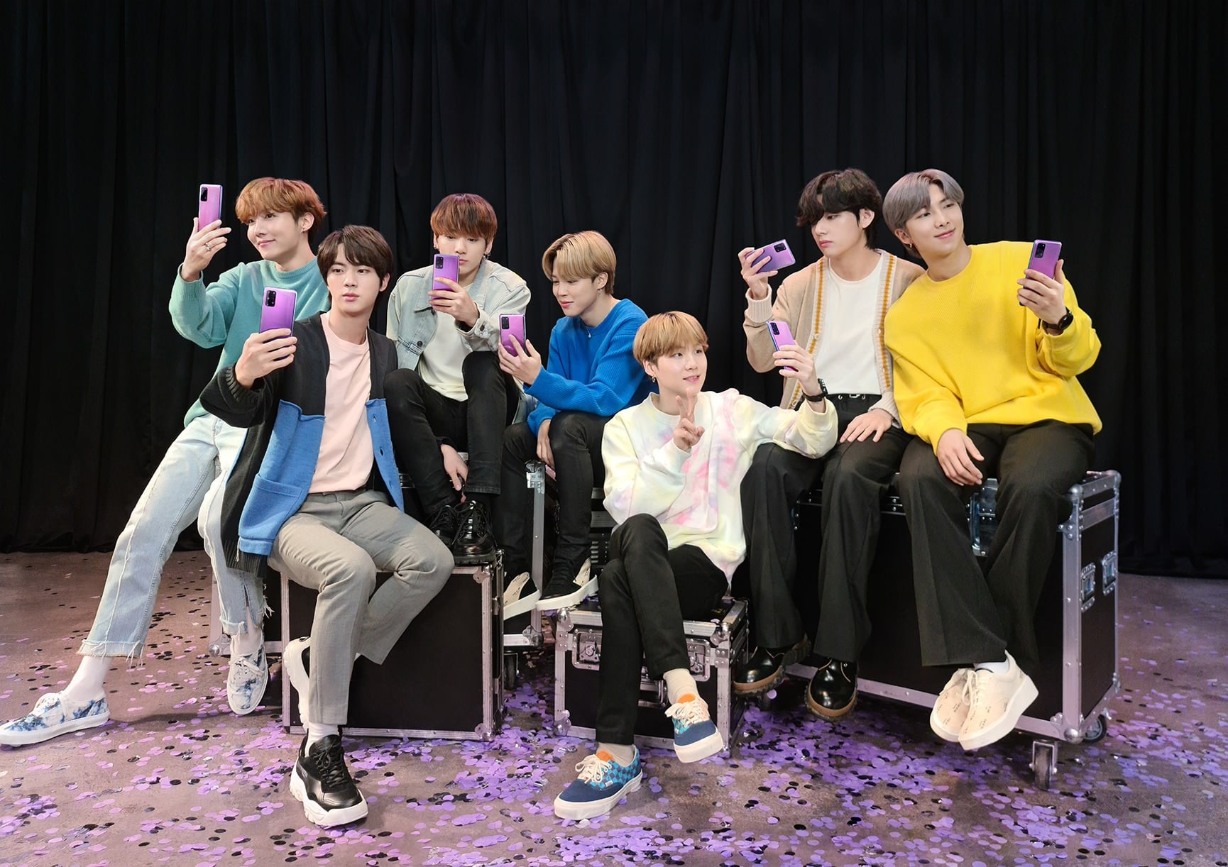 BTS防彈少年團坐在舞台上的輪箱上。每個人都拿著Galaxy S20+ 5G BTS Edition，地面上灑滿紫色的紙花。