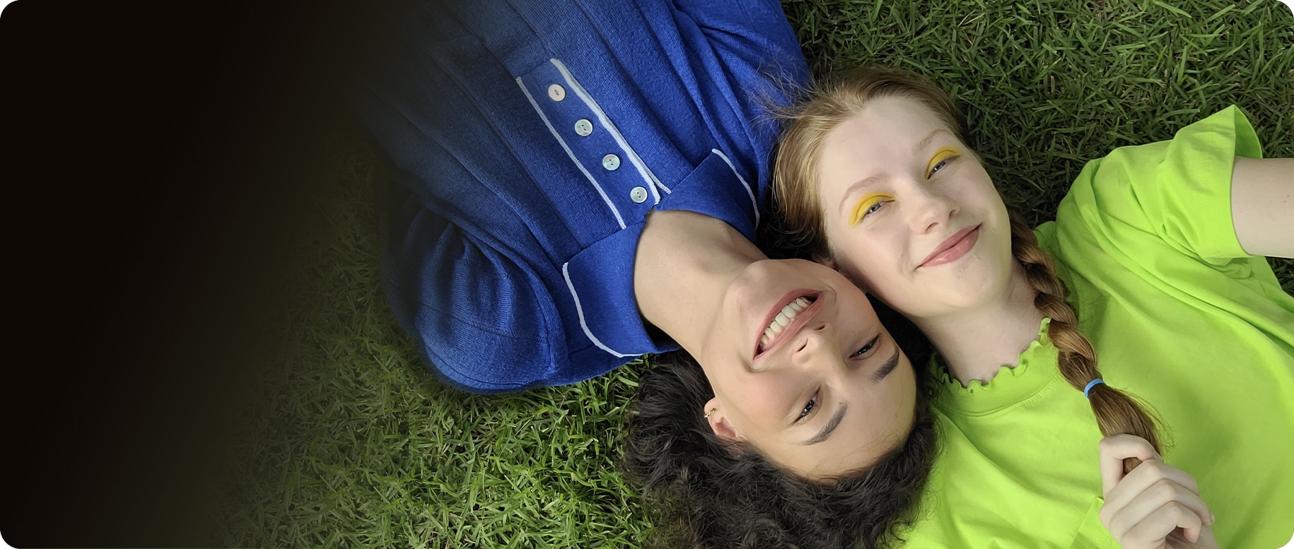 An aerial view of two women laying side-by-side on the grass. The woman on the left is wearing a blue collared shirt while the woman on the right is wearing a lime green top. Their heads are pressed together from opposite ends. They're both smiling at the camera.