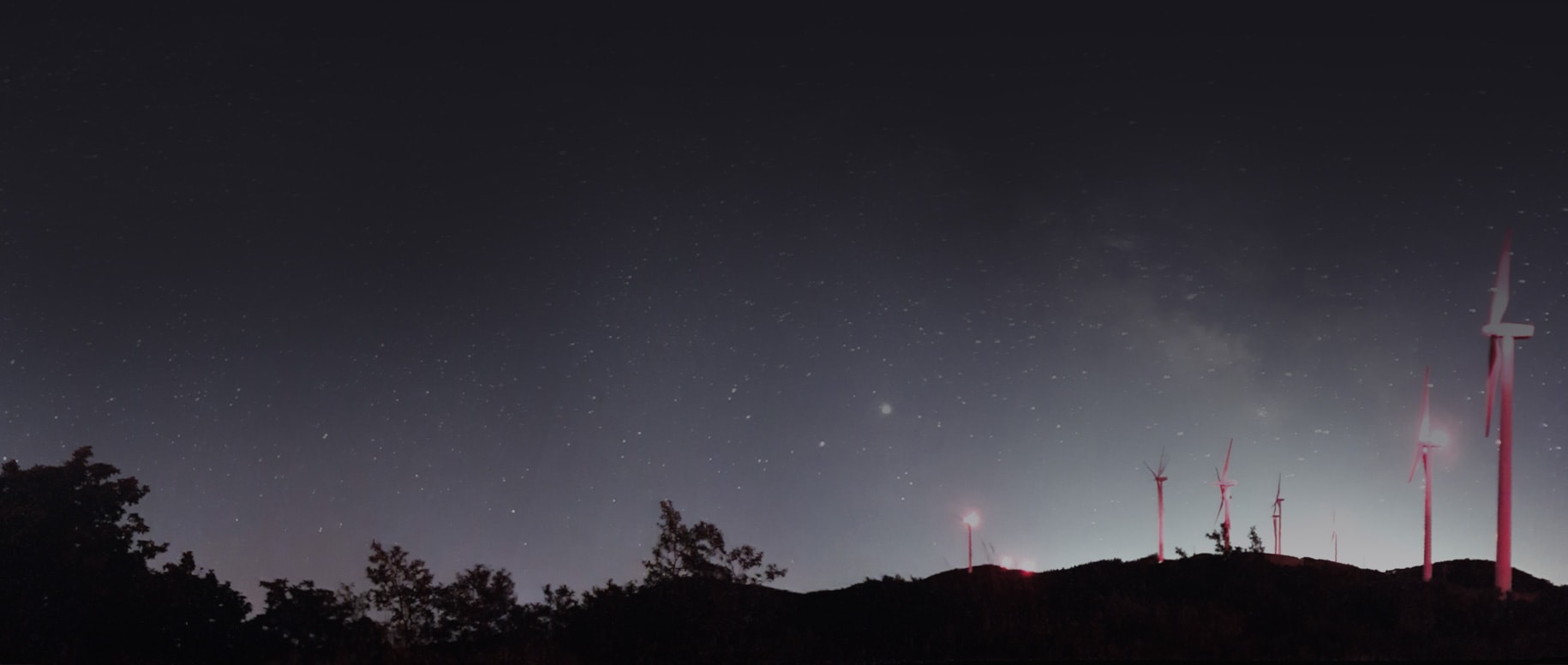 Una pequeña colina está salpicada de turbinas eólicas iluminadas en rojo en primer plano, con un cielo nocturno estrellado de fondo.