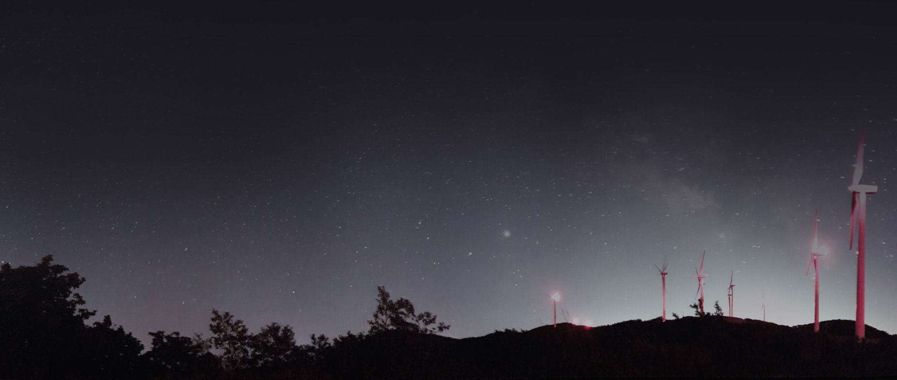 A small hill is dotted with red illuminated wind turbines in the foreground, with a sweeping starry night sky in the background.