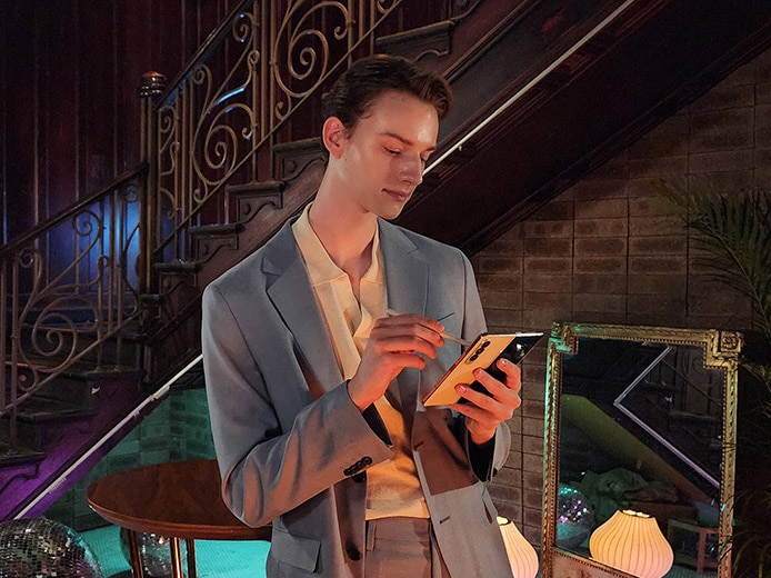 A man stands next to a stairwell in a dimly lit lounge, looking at the Main Screen of a Galaxy Z Fold4 in his hand. Nightography makes the colors bright and details of the subject and background clear even in a dark setting.