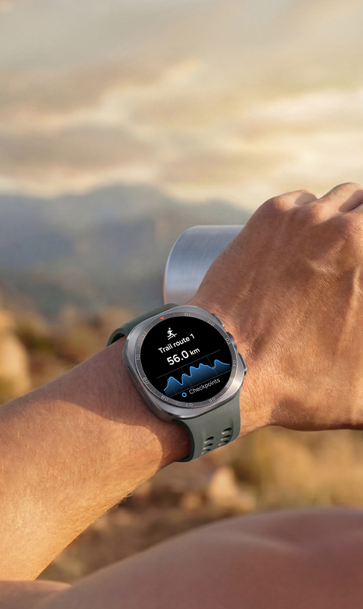 A person on a mountain trail with Galaxy Watch Ultra2 in Titanium Silver with a Marine Band in Olive on the wrist. Trail route and distance is displayed on screen.