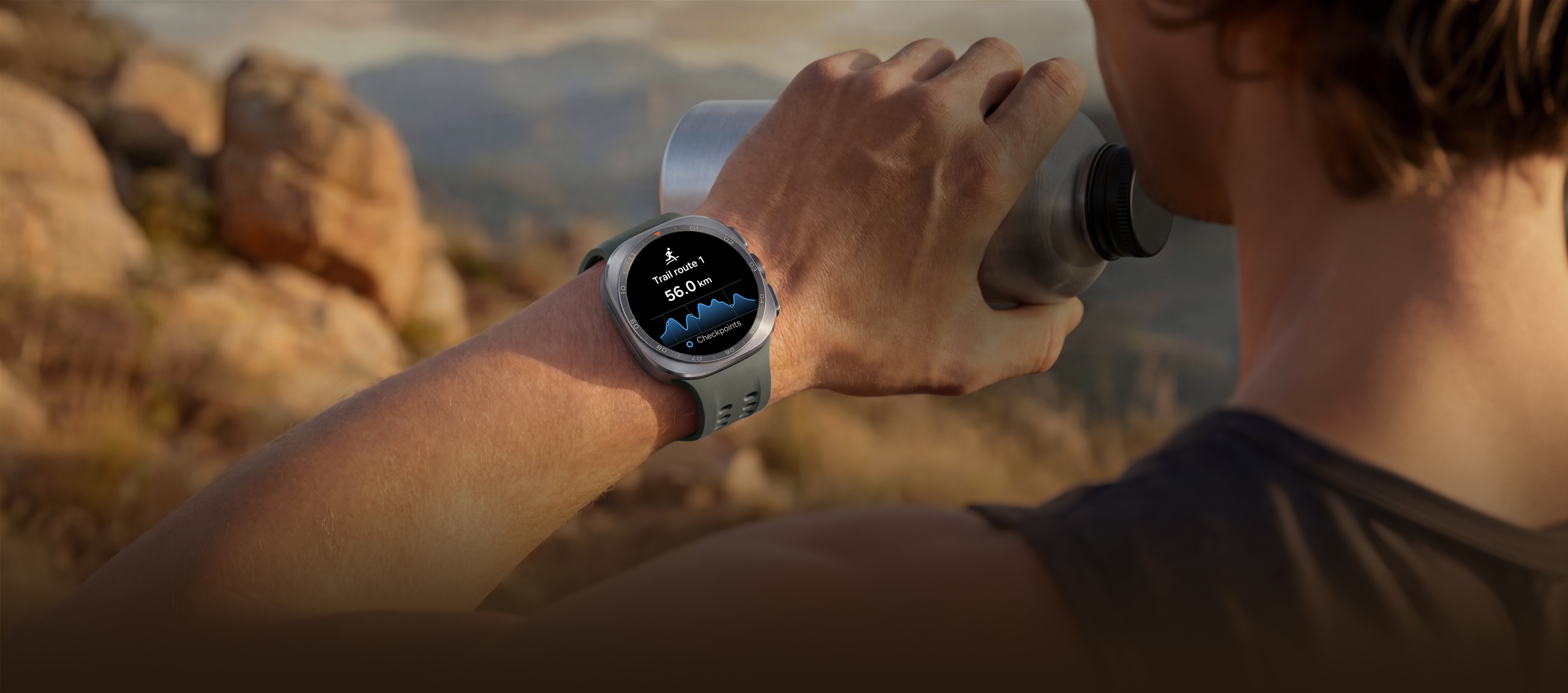 A man on a mountain trail with rocky terrain is holding a water tumbler in his hand while wearing a Galaxy Watch Ultra2 in Titanium Silver with a Marine Band in Olive, displaying a trail route with a distance, ideal for reliable guidance in challenging outdoor conditions during trail running and climbing.