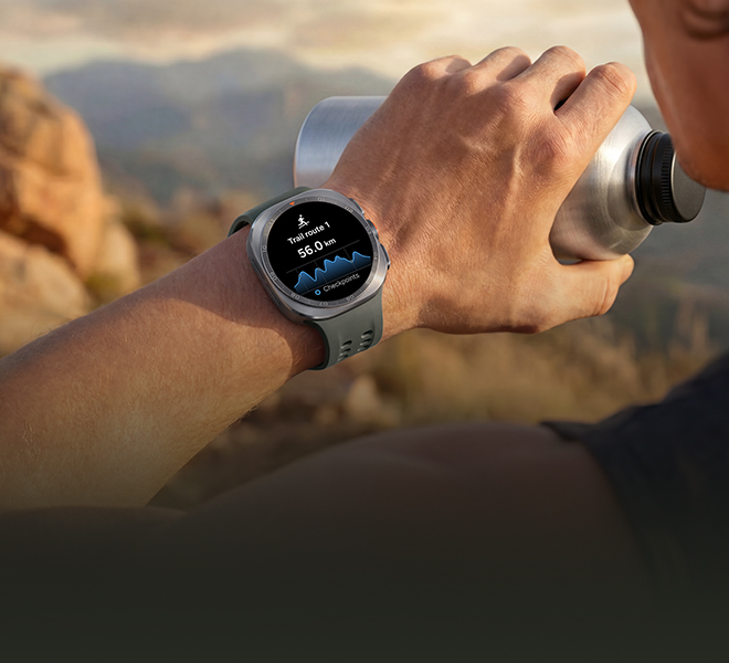A man on a mountain trail with rocky terrain is holding a water tumbler in his hand while wearing a Galaxy Watch Ultra2 in Titanium Silver with a Marine Band in Olive, displaying a trail route with a distance, ideal for reliable guidance in challenging outdoor conditions during trail running and climbing.