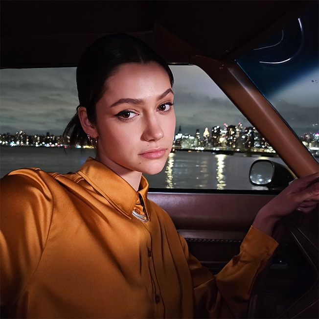 A night time selfie taken in a car with a waterfront city skyline in the background. The color …