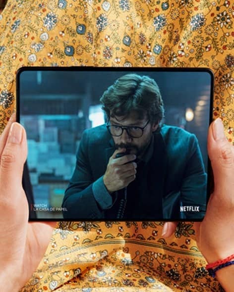 Hai bàn tay cầm chiếc điện thoại Galaxy Z Fold3 5G, mở ra. Màn hình chính hiển thị cảnh từ một video Netflix với hình ảnh một chàng trai đang giơ tay lên mặt.