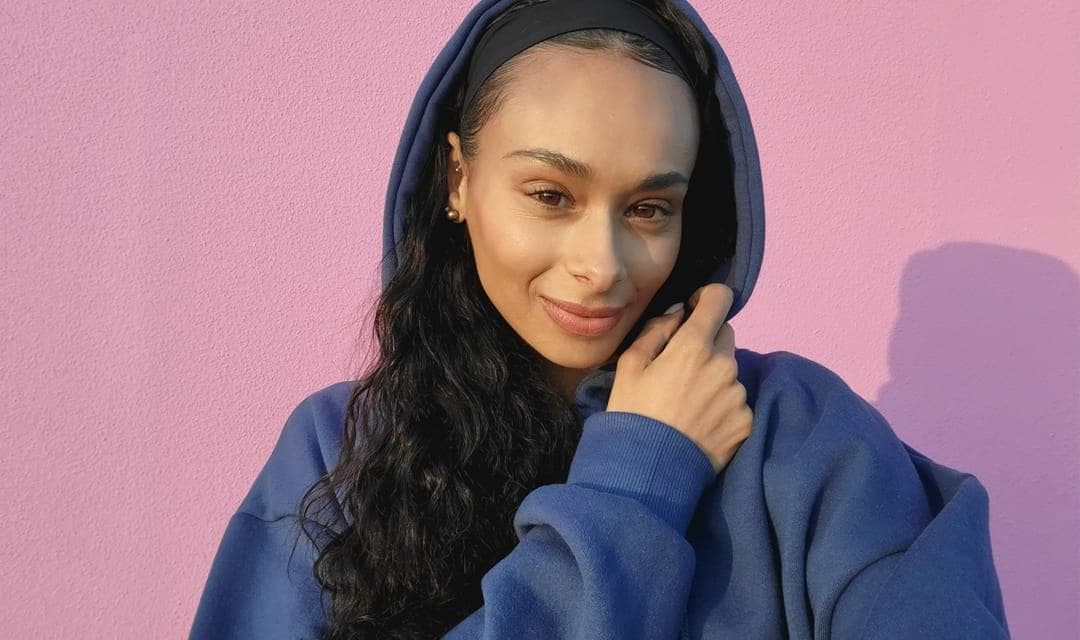 Woman smiling as she takes a selfie against a pink wall.